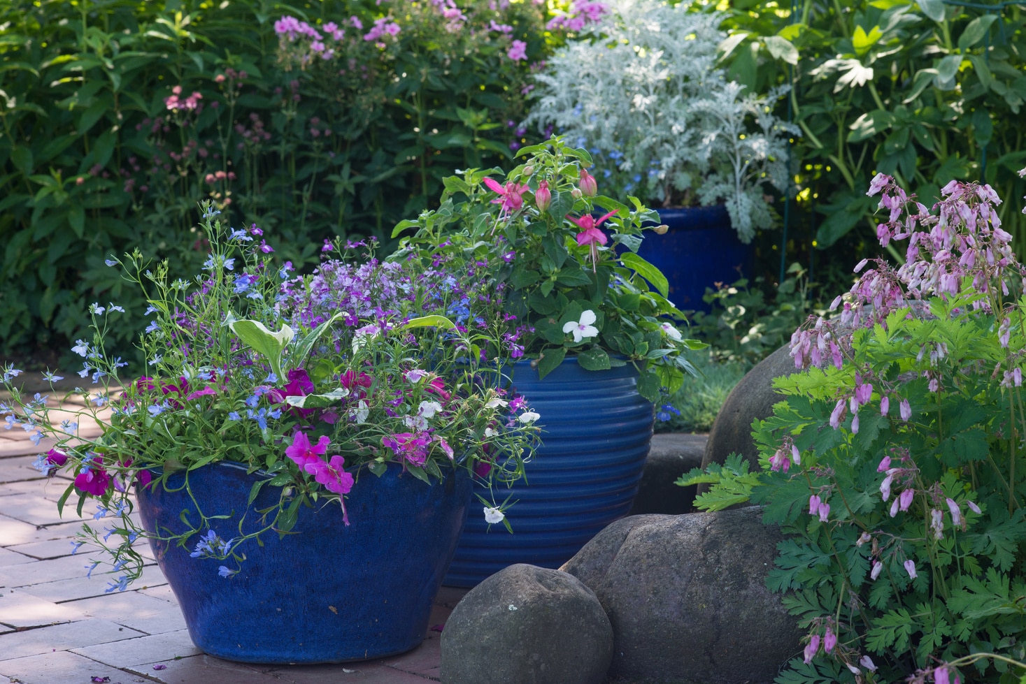 Bright blue pots overflow with colorful flowers in a shaded brick patio.