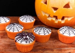Halloween themed cupcakes with a black spider web iced over white icing, held in orange cupcake cases.
