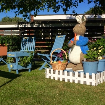 Large knitted Peter Rabbit eating basket of produce next to plant pots