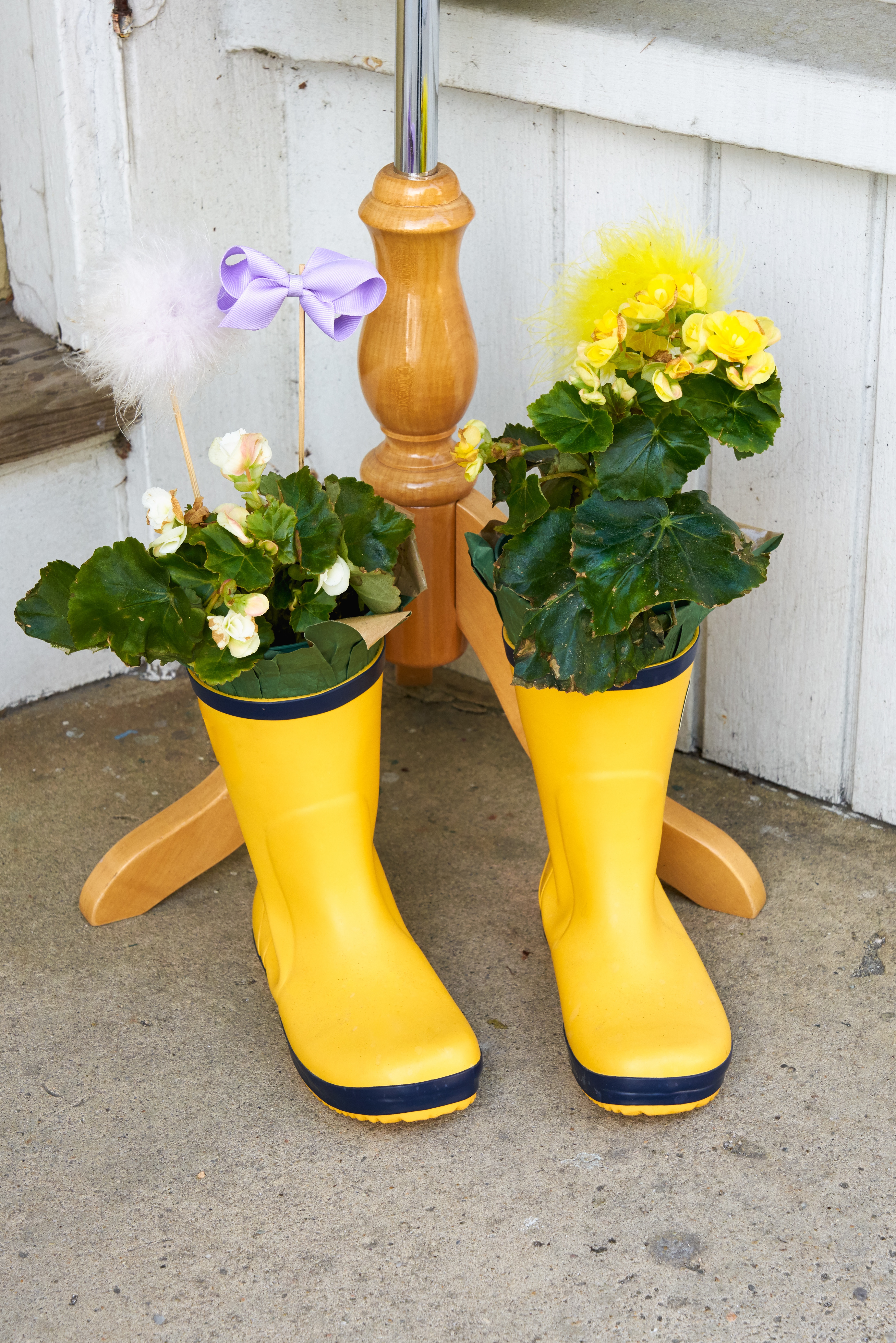 childs bright yellow rubber rain boots used as flower planters sitting on the sidewalk with a lavender ribbon decoration