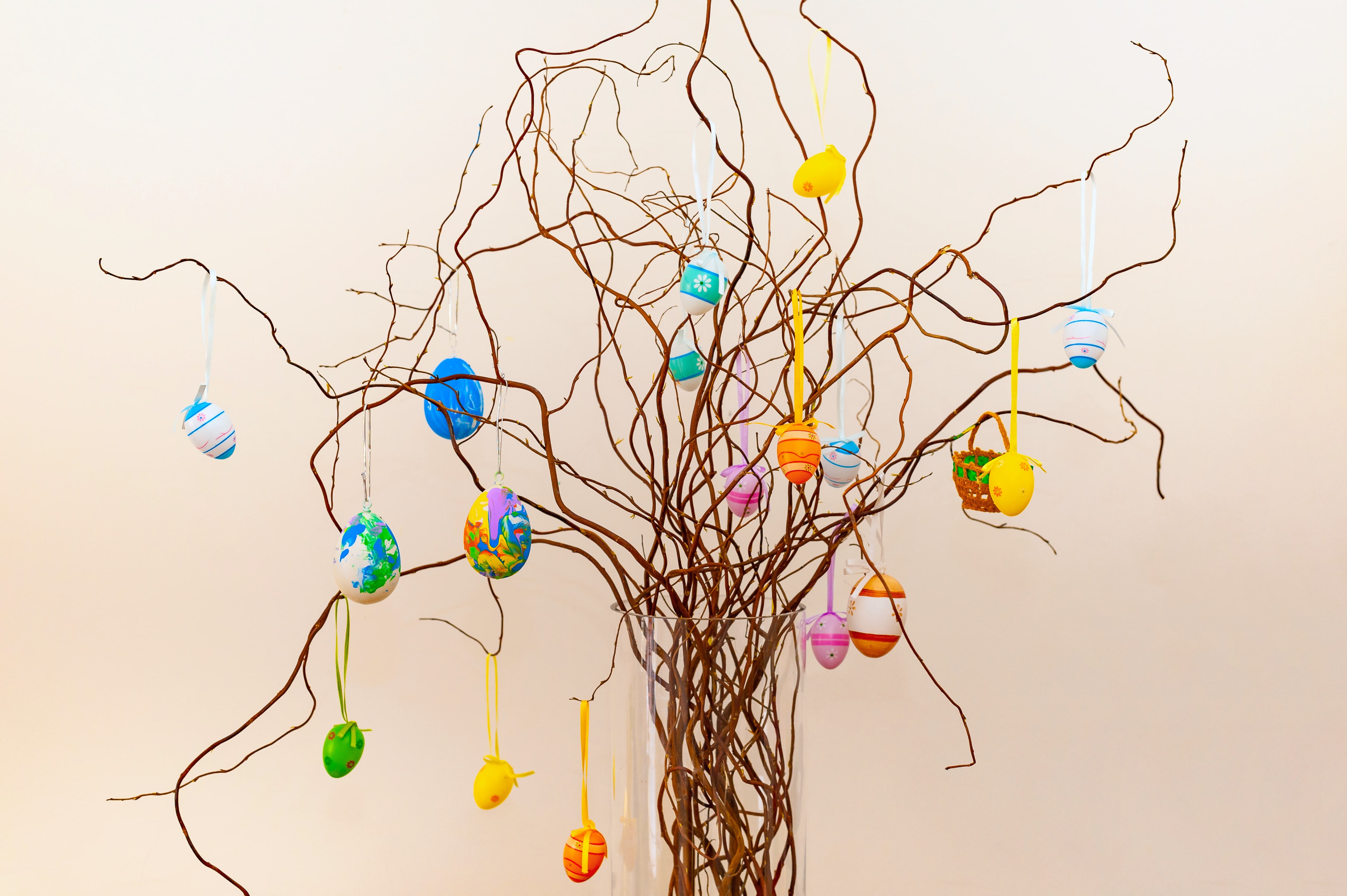 Easter decoration made of rustic twigs and colourful eggs hanging on ribbons in tall glass vase on neutral creamy backgroud.