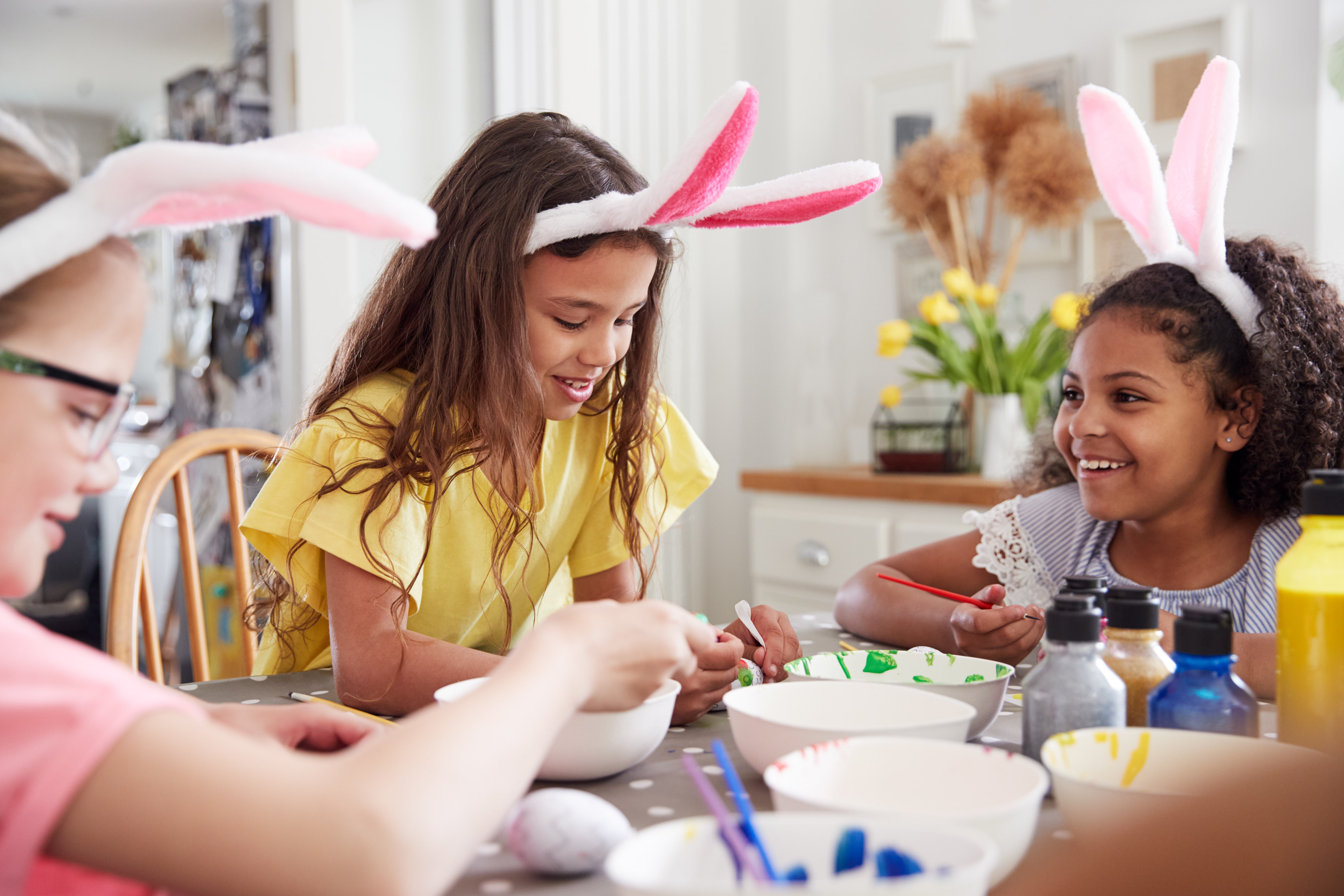 Three Girls Wearing Bunny Ears Sitting At Table Decorating Eggs For Easter At Home