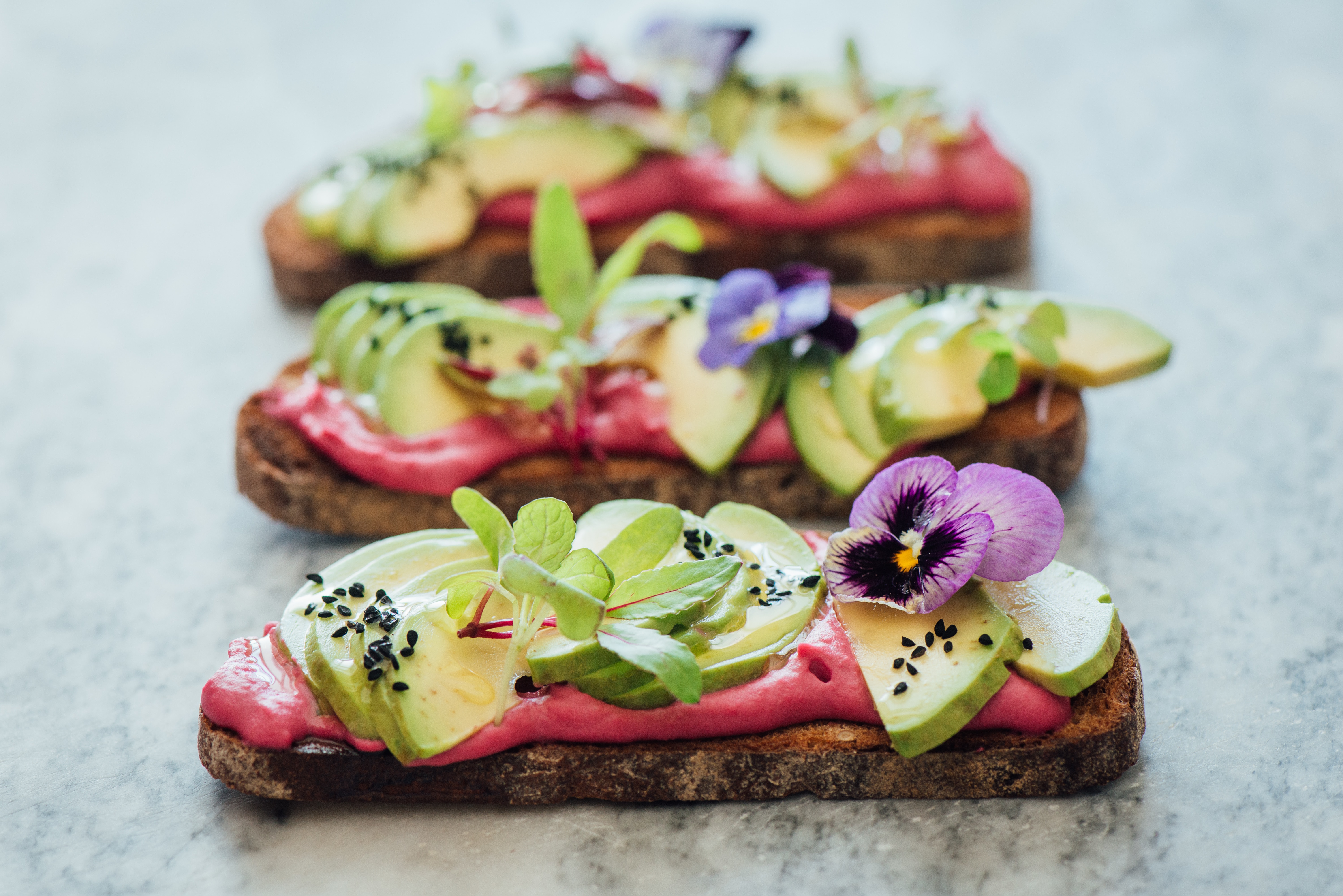 Low angled shot of avocado rye toast on beetroot hummus with edible purple pansy flowers, micro greens and nigella seeds or onion seeds, on marble background. soft focused background