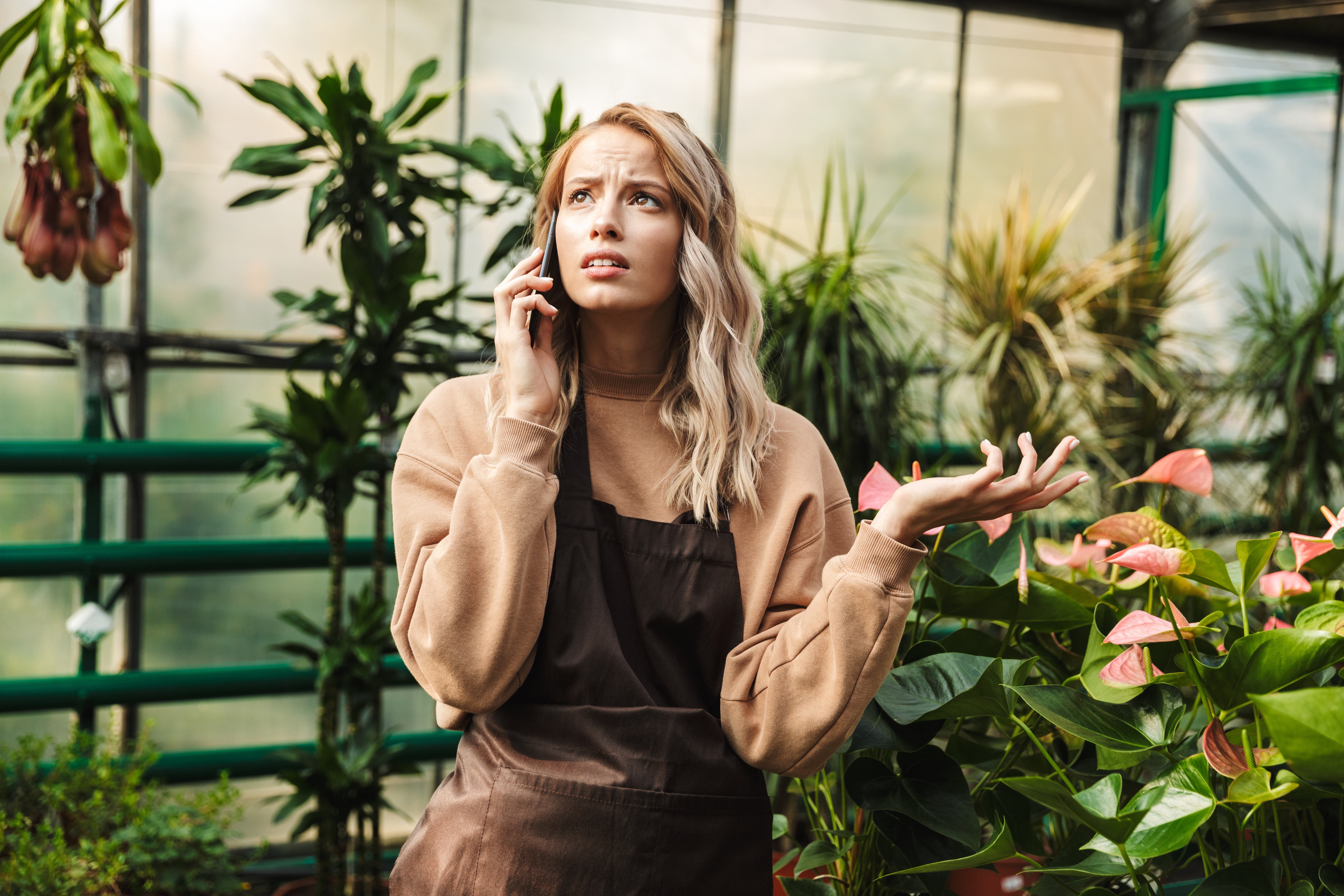 Image of beautiful confused sad gardener woman posing in the nature greenhouse garden talking by mobile phone.
