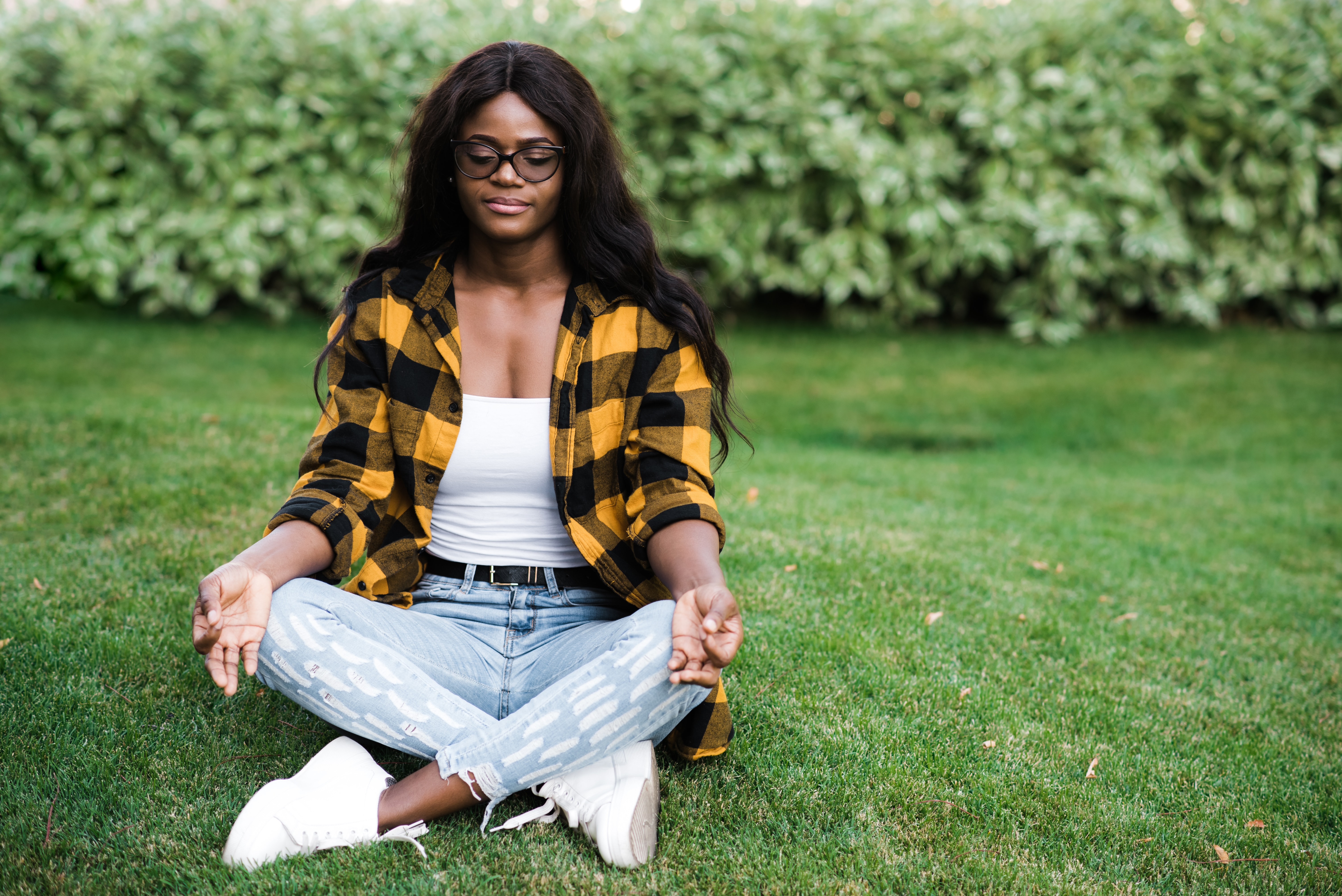 Young African American woman dressed in stylish casual clothes meditates in the park while sitting on the grass in lotus position with closed eyes