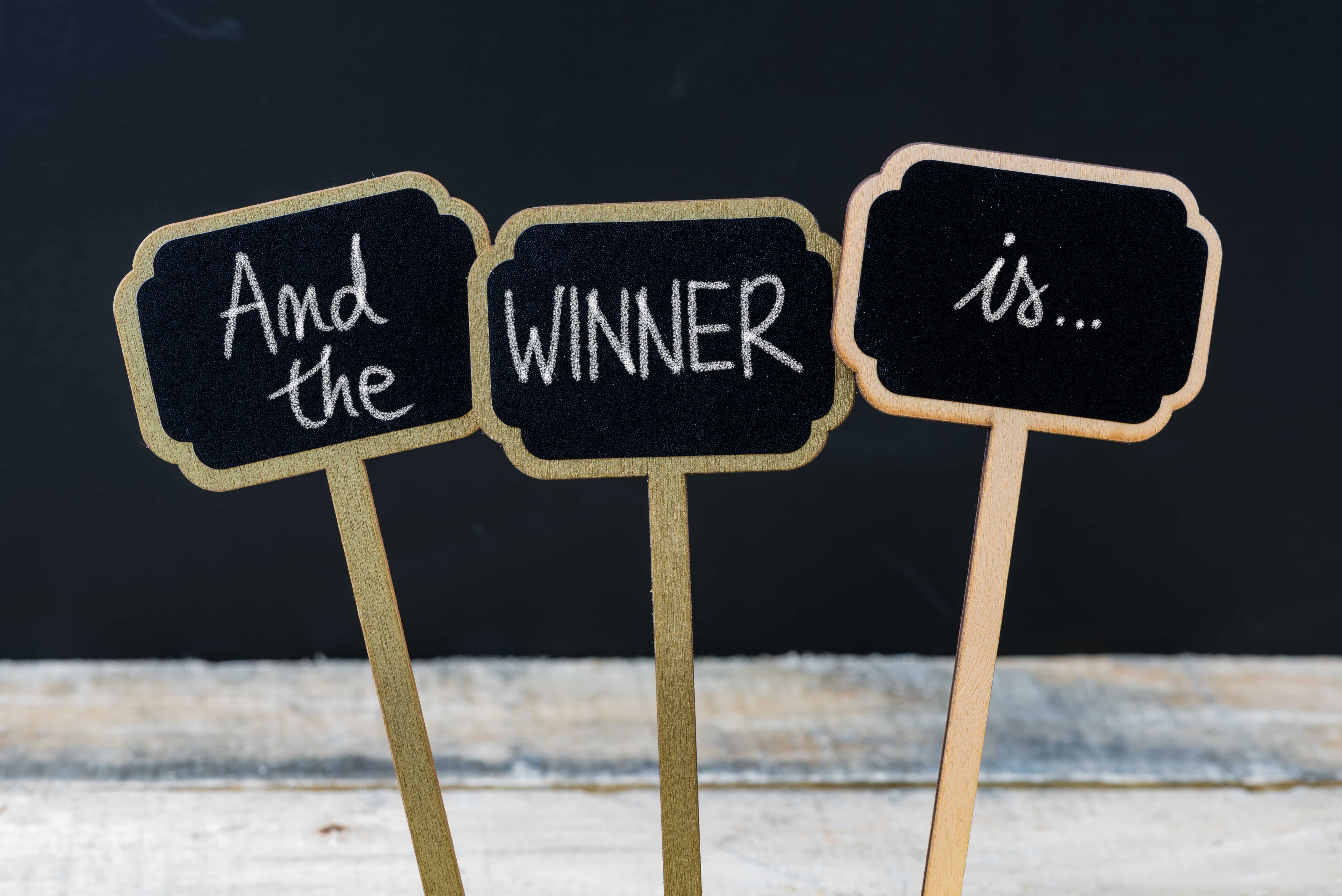 Motivational message And The Winner is... written with chalk on wooden mini blackboard labels, defocused chalkboard and wood table in background